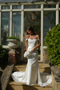 Bride in a white off the shoulder wedding dress featuring fit and flare silhouette and draped detail standing in a greenhouse-like setting with greenery.