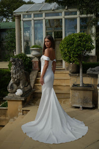 Woman in a white off-shoulder dress standing on steps with a greenhouse in the background