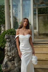 Bride in a white off the shoulder fitted satin  gown holding a bouquet standing next to a stone lion sculpture outdoors.