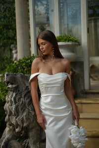 Bride in a white off the shoulder fitted wedding dress holding flowers, standing next to a stone lion sculpture.
