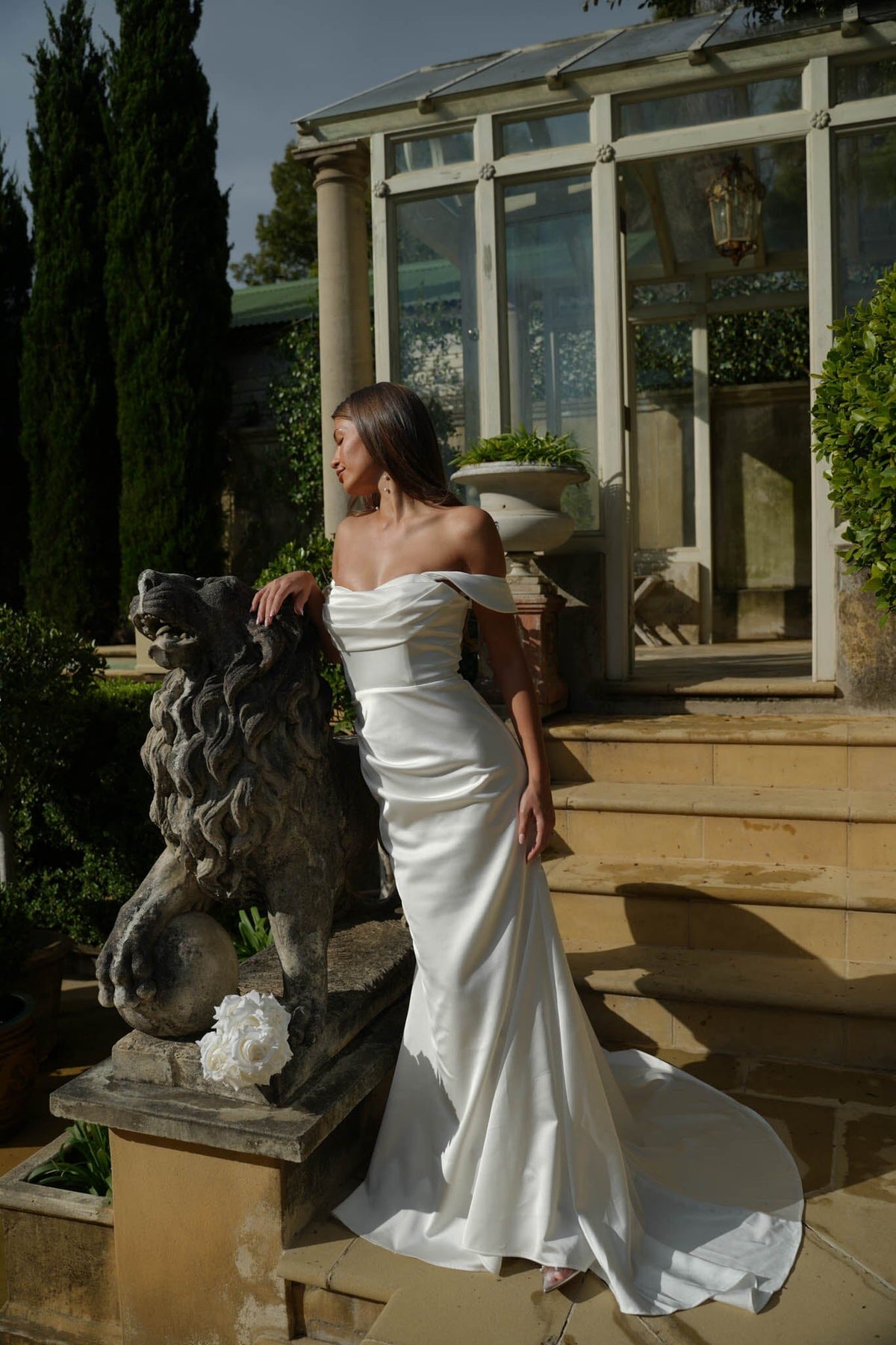 Woman in a white off-shoulder satin wedding gown standing next to a stone lion statue in an outdoor setting.