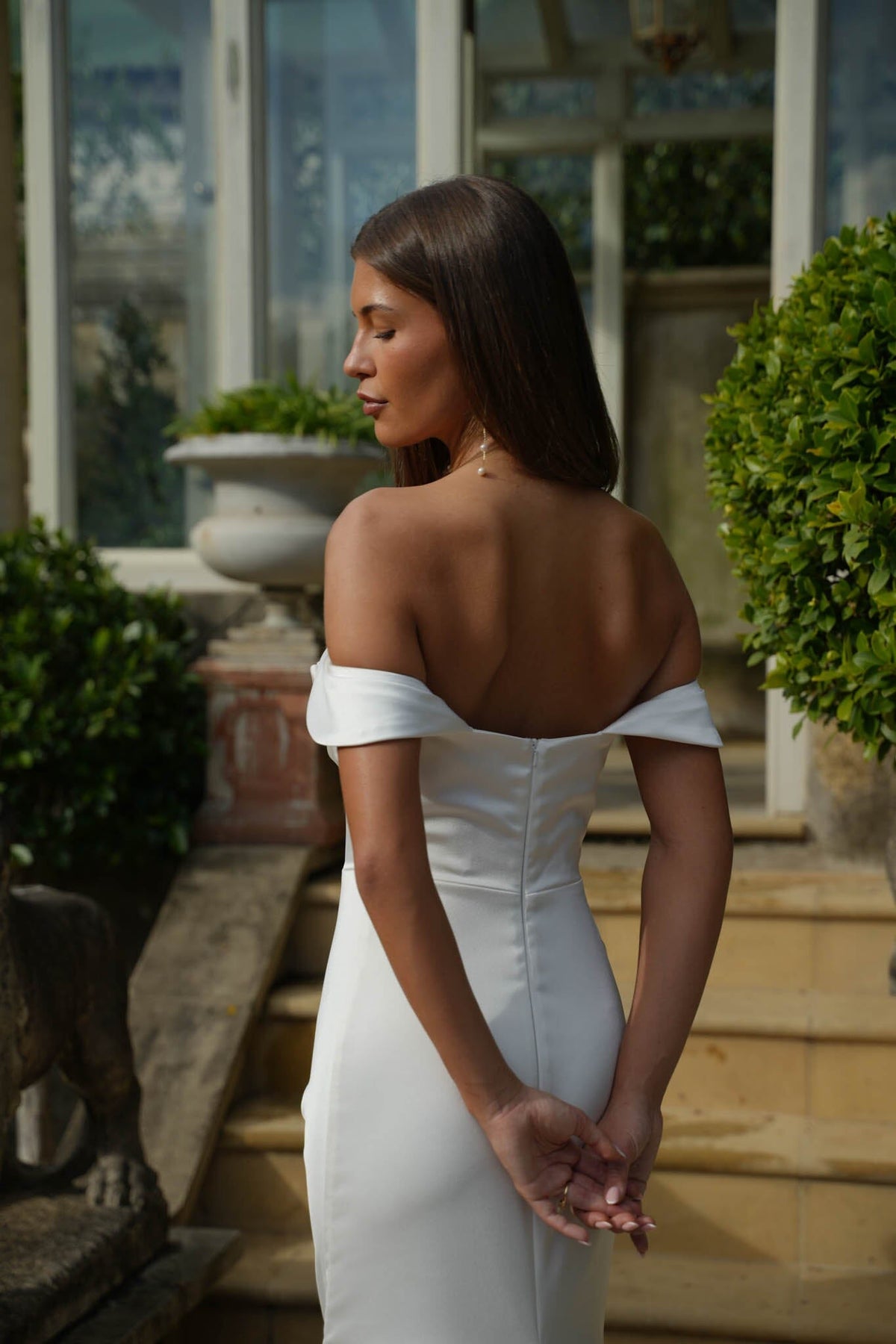 Woman in a white off-shoulder dress standing outdoors with greenery and architectural elements in the background.