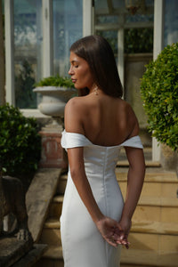 Woman in a white off-shoulder dress standing outdoors with greenery and architectural elements in the background.