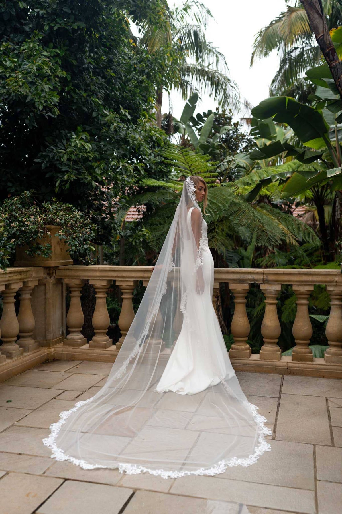 A bride in a white wedding gown with a long sheer veil with lace edging detail stands on a stone terrace surrounded by lush tropical greenery.