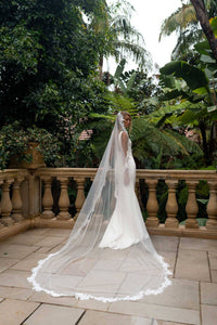 A bride in a white wedding gown with a long sheer veil with lace edging detail stands on a stone terrace surrounded by lush tropical greenery.