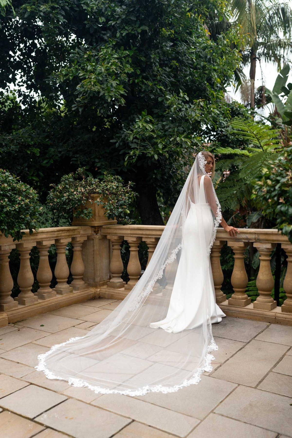 A bride in a white wedding gown with a long lace-trimmed veil stands on a stone terrace, leaning on a balustrade with lush green garden foliage in the background.