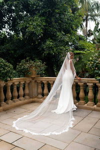 A bride in a white wedding gown with a long lace-trimmed veil stands on a stone terrace, leaning on a balustrade with lush green garden foliage in the background.