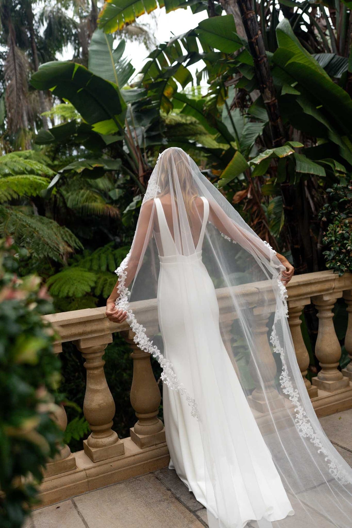 A bride in a white wedding gown and long sheer veil with floral lace trim stands on a stone balcony surrounded by lush tropical greenery, viewed from behind. 