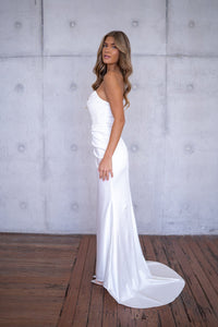 Side image of white bustier strapless fitted stretch satin wedding gown with draped pleats and high side slit
