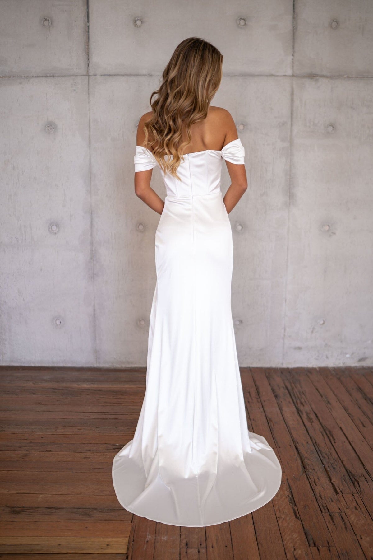Woman wearing a white off-shoulder wedding dress against a plain concrete wall