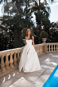 Modern bride wearing white strapless ballgown wedding dress with a Basque waist, draped neckline and leg slit walking along poolside