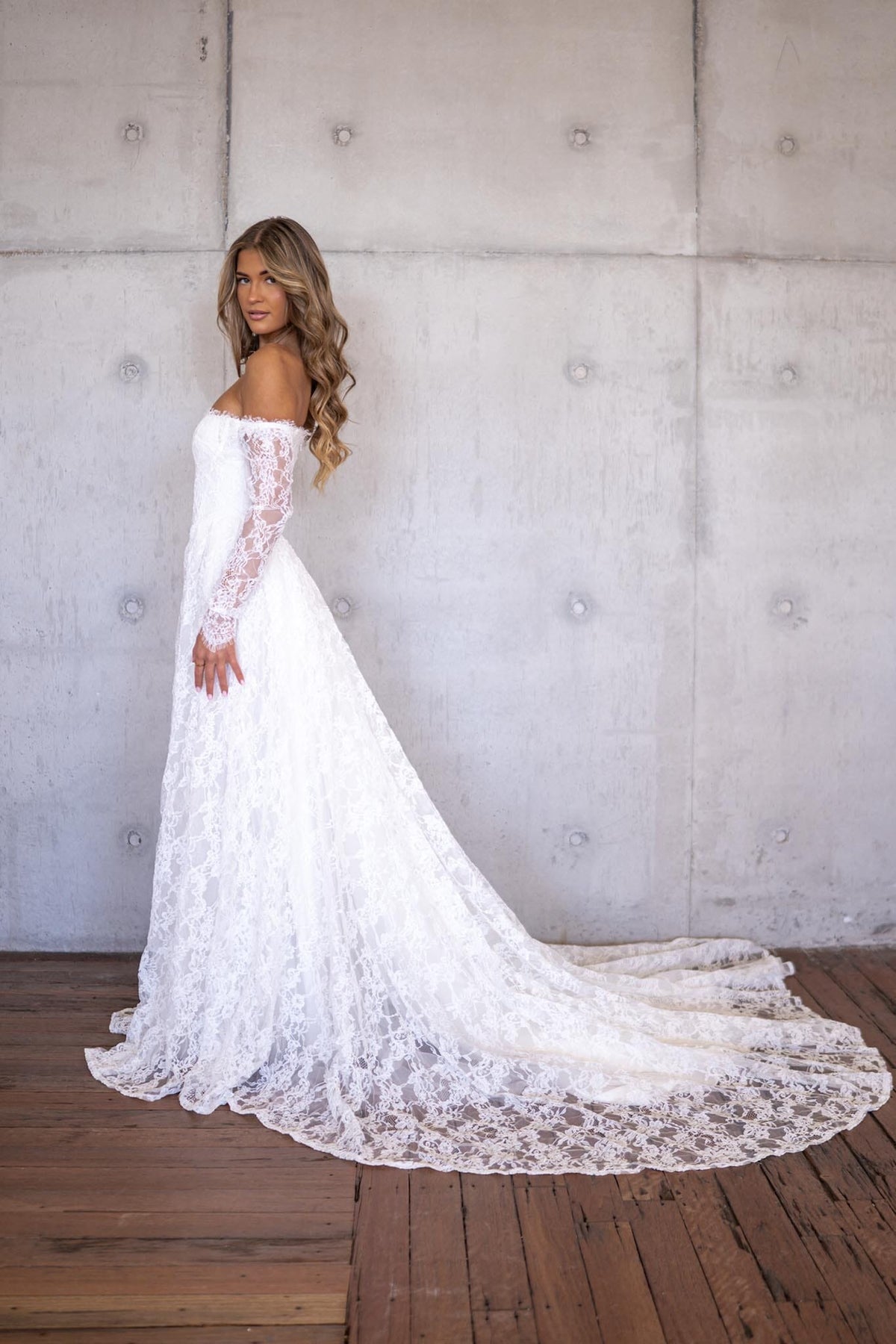 Side Image of Ivory White Off The Shoulder Long Sleeve Lace Wedding Gown featuring Sweetheart Neckline with Lace Trimming Detail, Sheer Long Sleeves, Corset Bodice, Basque Waistline, Full A-line Skirt and Long Train.