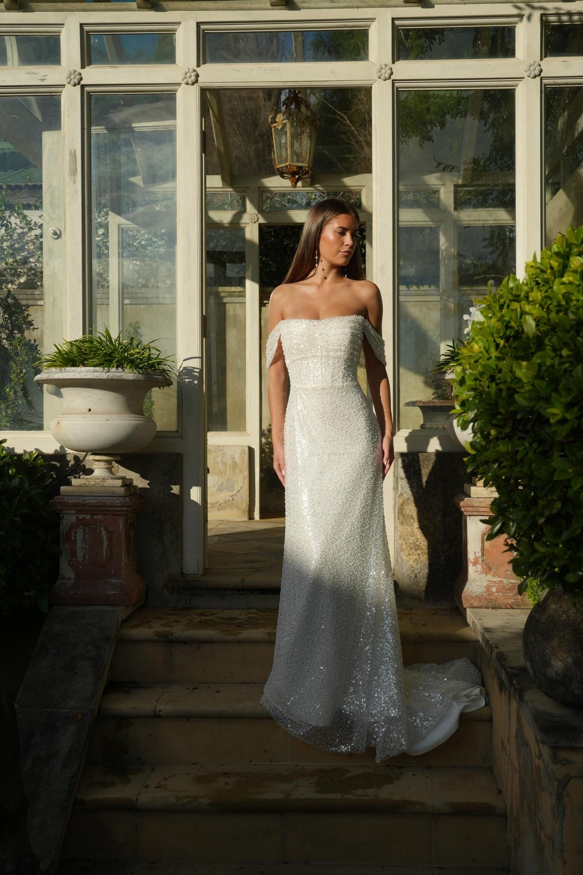 Bride in a white off-shoulder beaded wedding gown standing on stone steps with a glasshouse background