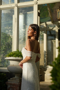Bride in a white off-shoulder wedding dress standing in a sunlit room with large windows and greenery.