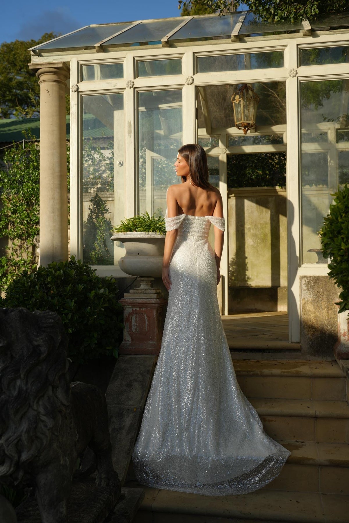 Woman in a white off-shoulder sparkly wedding dress standing on steps in front of a glasshouse.