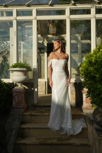 Bride in a white off-shoulder beaded wedding gown standing in a sunlit greenhouse.
