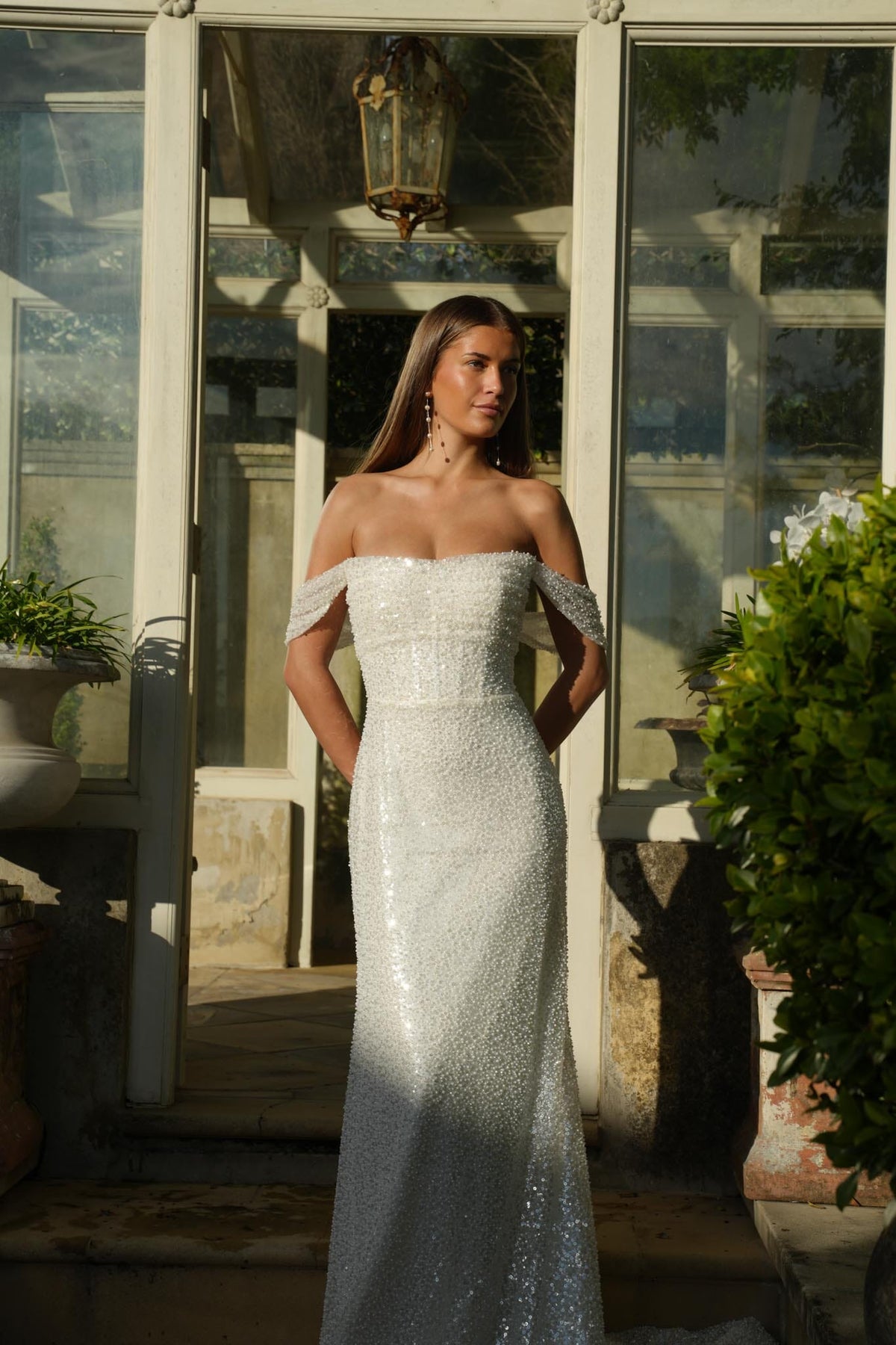 Woman in a sparkling off-shoulder wedding gown standing outdoors with reflective surfaces and plants.