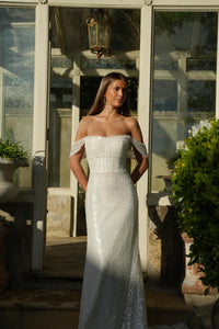Woman in a sparkling off-shoulder wedding gown standing outdoors with reflective surfaces and plants.