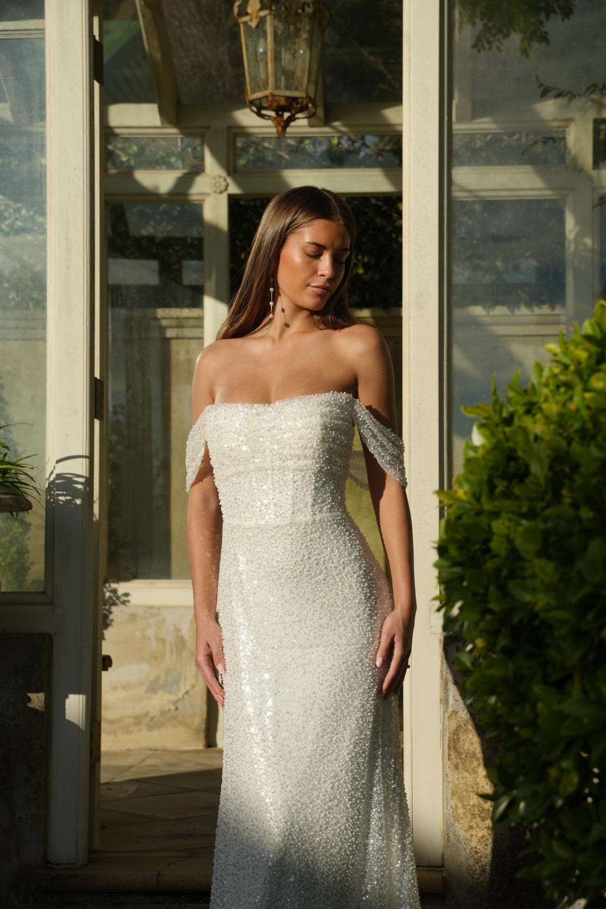 Bride in a sparkly off-shoulder wedding gown standing outdoors near a building entrance.